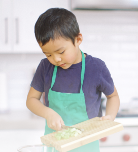 A young boy in a blue t-shirt and green aprons scrapes the vegetables he chopped off a cutting board and into a bowl.